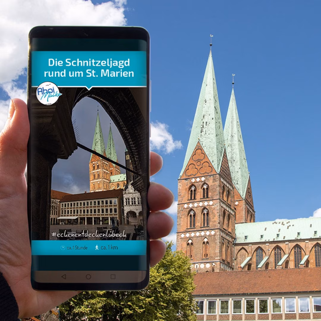Smartphone zeigt Schnitzeljagd rund um St. Marien in Lübeck, im Hintergrund die Kirche mit grünem Doppelturm.