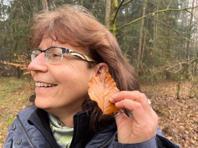 Eine Frau hält bei einem Waldbaden-Retreat ein Blatt an ihr Ohr und lauscht dem Knistern
