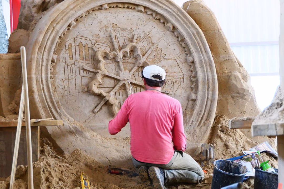 Sandskulpturenausstellung: Künstler kniet und bearbeitet ein rundes Reliefmedaillon mit Stadtmotiven aus Sand.