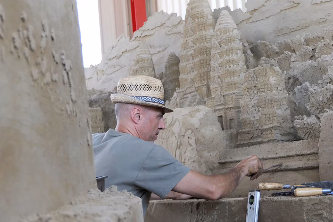 Sandskulpturenausstellung: Mann mit Strohhut formt filigrane Tortürme einer Tempelanlage aus verdichtetem Sand.
