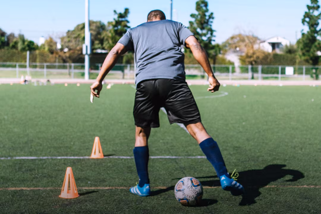 Ein Fußballtrainer läuft auf Rasen mit einem Fußball um orangefarbene Hütchen herum, den Rücken zur Kamera gewandt.