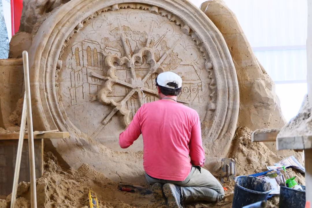Sandskulpturenausstellung: Künstler kniet und bearbeitet ein rundes Reliefmedaillon mit Stadtmotiven aus Sand.