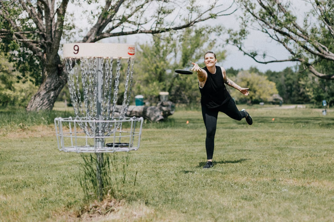 Frau in schwarzer Sportkleidung wirft beim Disc Golf eine Scheibe auf den Korb auf einer Grünfläche.