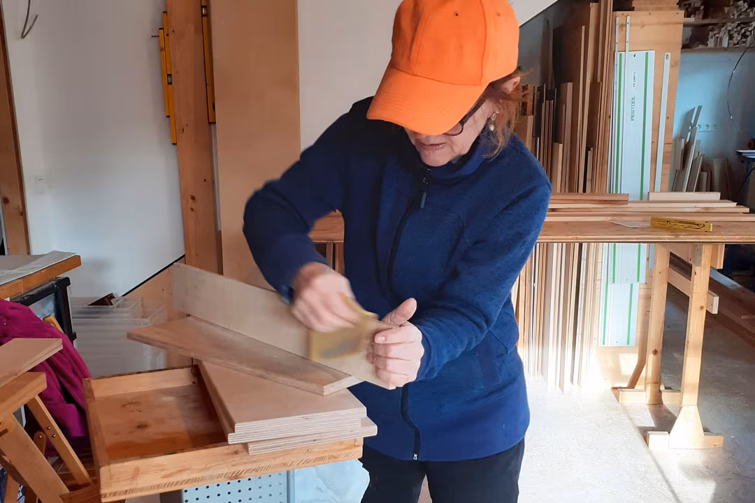 Frau beim Mädelsabend in der Holzwerkstatt: Sie trägt Kappe und schleift konzentriert ein Holzbrett für ihr eigenes Unikat.