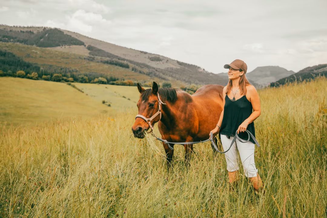 Eine Frau mit Cap führt ein Quarter Horse auf einer Wanderung durch Wiesen, im Hintergrund sanfte Hügel.