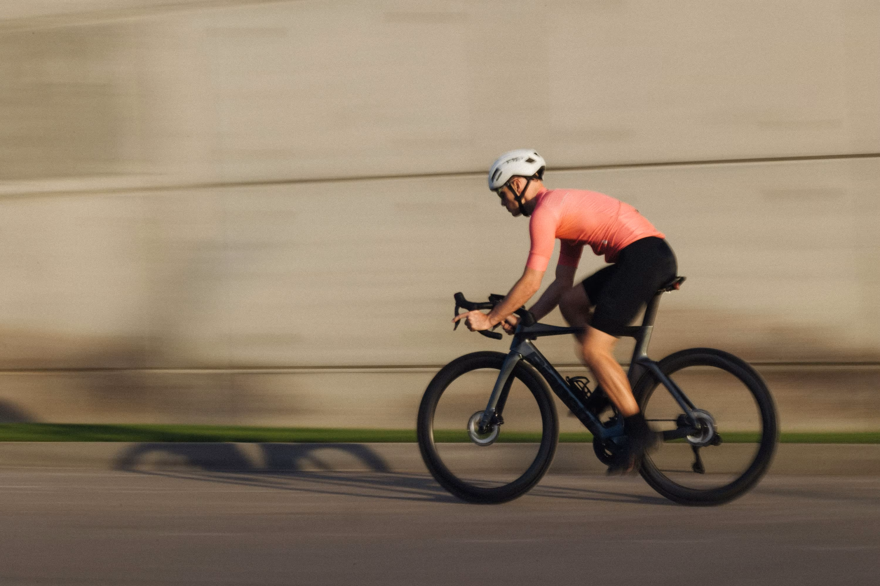 Rennradfahrer auf schwarzem Rennrad fährt schnell auf einer Straße, im Hintergrund eine Betwonwand.