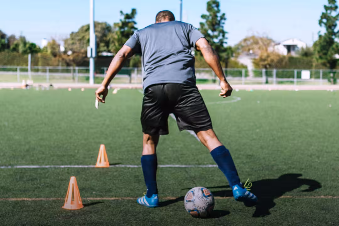 Ein Fußballtrainer läuft auf Rasen mit einem Fußball um orangefarbene Hütchen herum, den Rücken zur Kamera gewandt.