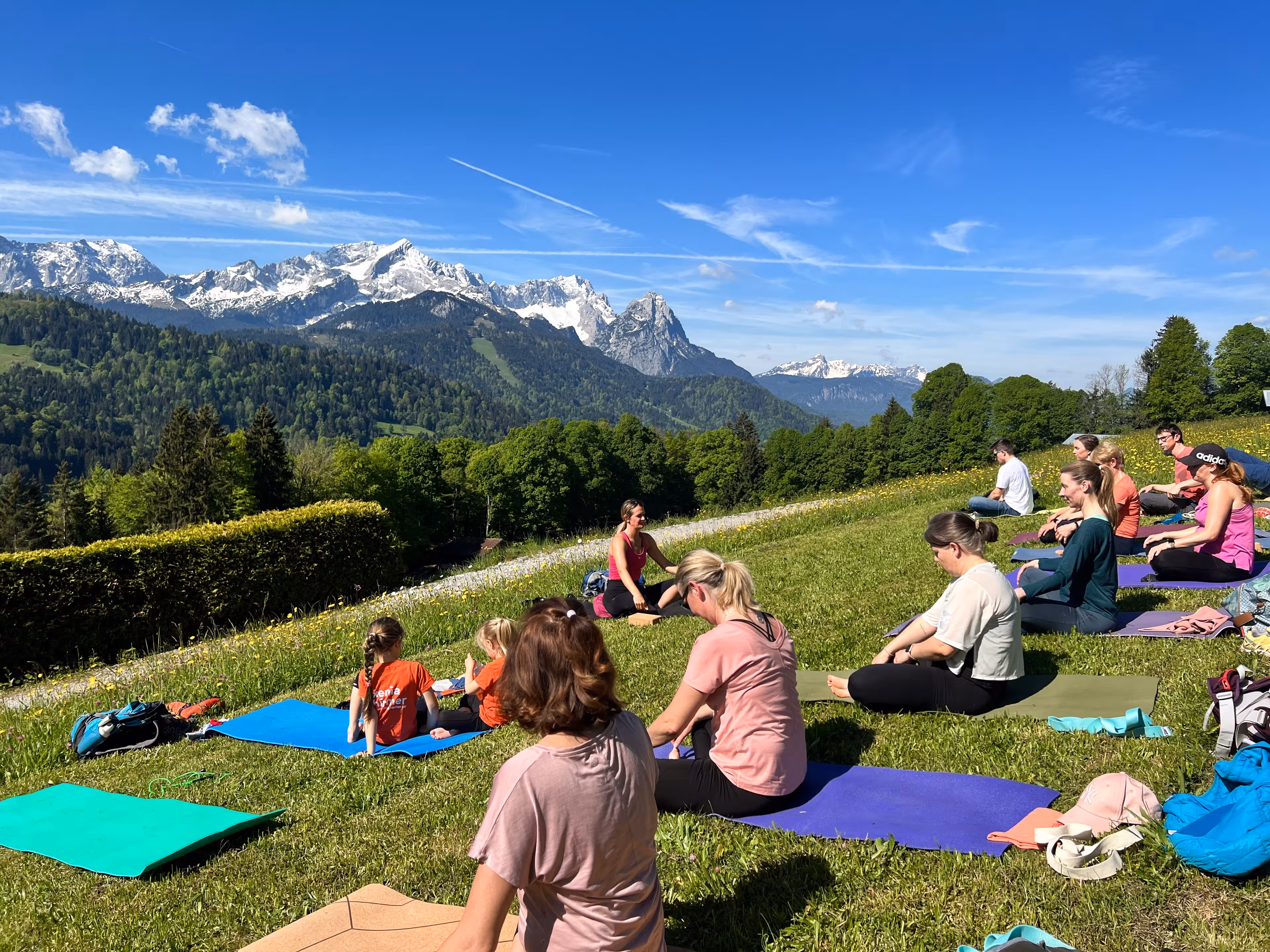 Teilnehmende sitzen beim Yoga in den Bergen im Schneidersitz, im Hintergrund sind die Alpen zu sehen