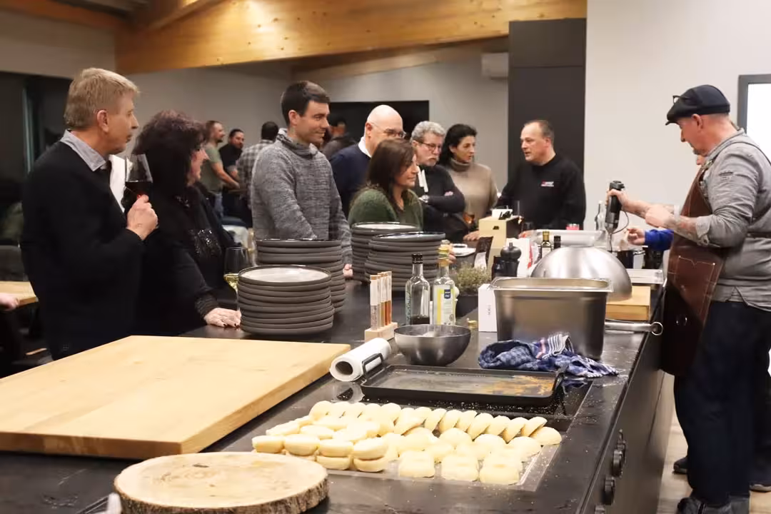 Mehrere Menschen stehen in der Eventküche beim Afterwork Burger-Grillkurs und schauen dem Profikoch zu. Im Vordergrund Buns.