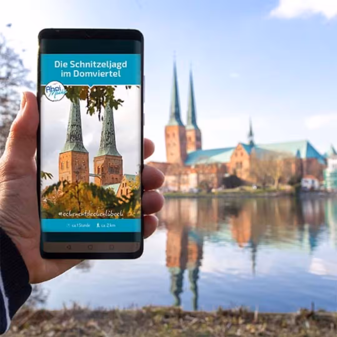 Smartphone zeigt Schnitzeljagd im Domviertel in Lübeck, im Hintergrund spiegeln sich die Domtürme im Wasser.