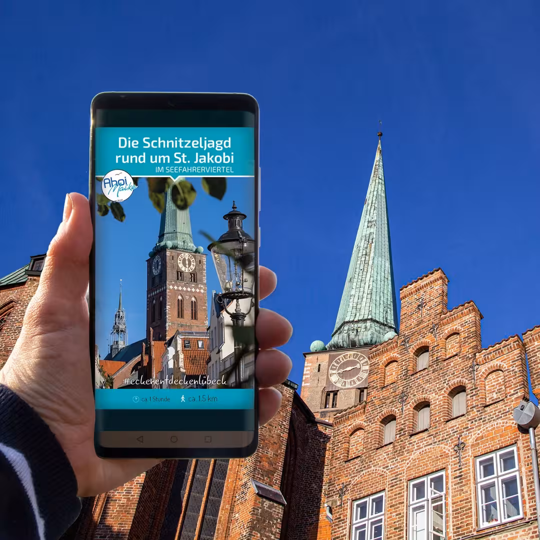 Smartphone zeigt Schnitzeljagd rund um St. Jakobi in Lübeck, im Hintergrund die Kirche mit grünem Turm.