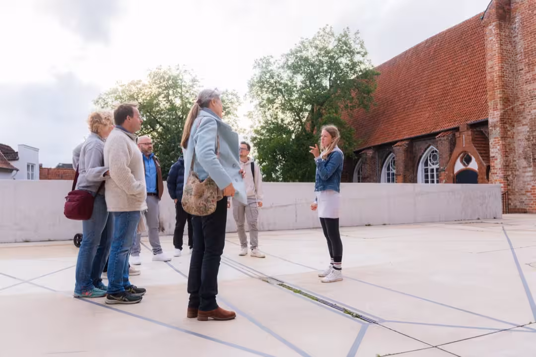 Eine Stadtführerin steht auf einem hellen Kirchenvorplatz und redet mit der vor ihr stehenden Gruppe.
