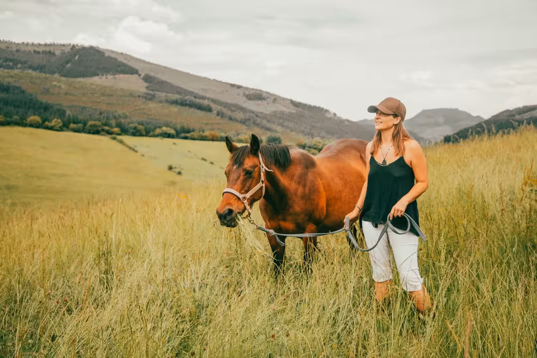 Eine Frau mit Cap führt ein Quarter Horse auf einer Wanderung durch Wiesen, im Hintergrund sanfte Hügel.