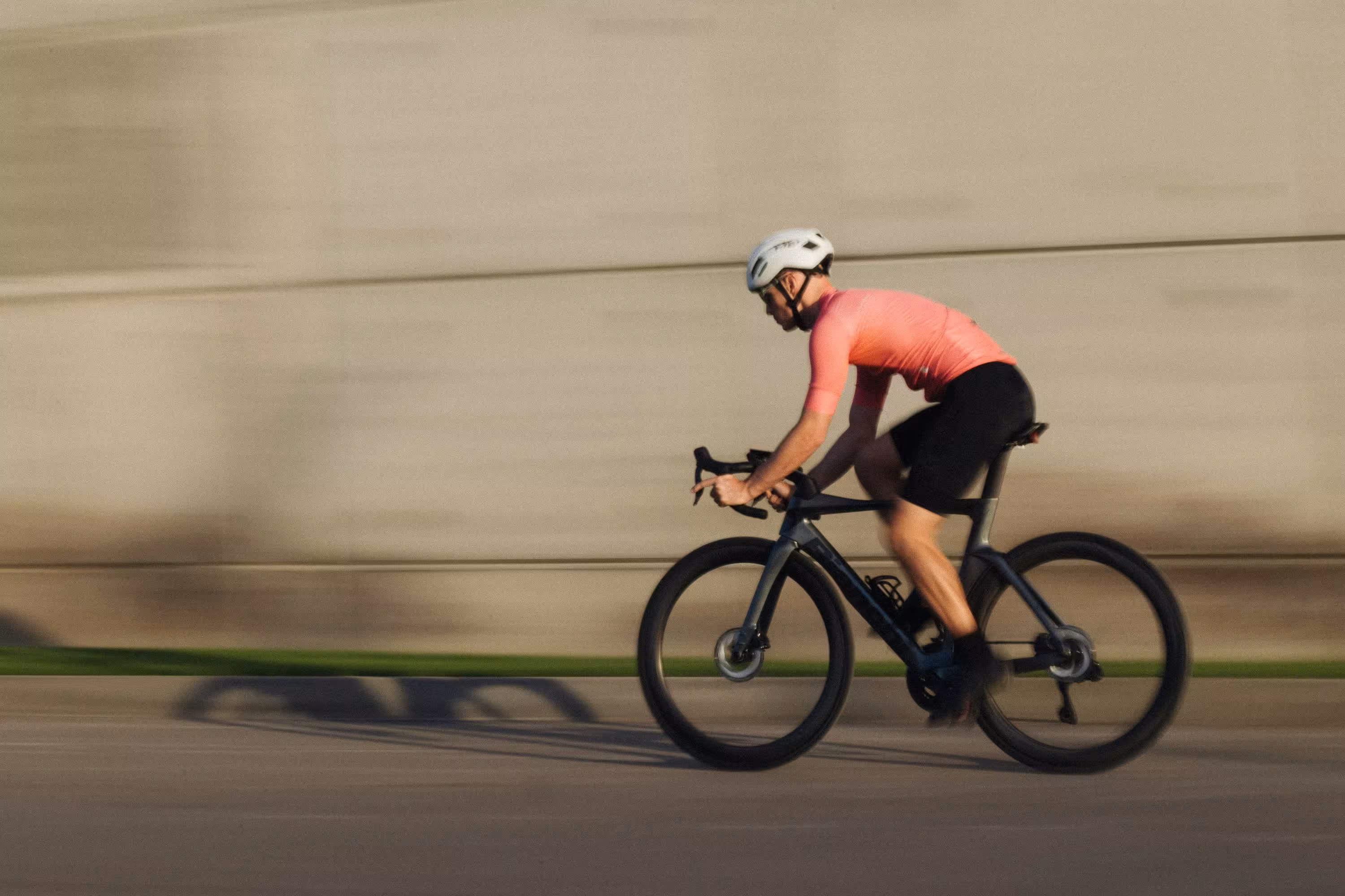 Rennradfahrer auf schwarzem Rennrad fährt schnell auf einer Straße, im Hintergrund eine Betwonwand.