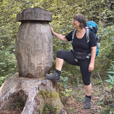 Frau mit Rucksack wandert im Wald und posiert lachend neben einer großen hölzernen Skulptur in Form eines Pilzes.