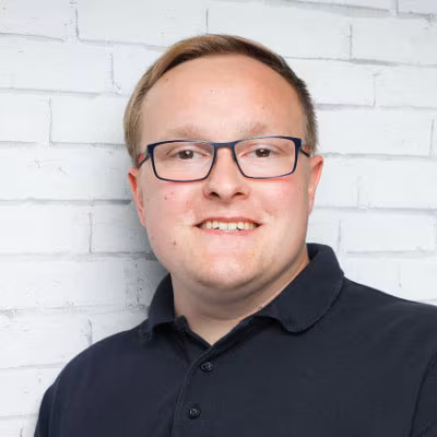Ein Porträtfoto von Marcel Bodien, Fußballtrainer mit schwarzer Brille und schwarzem Polo-T-shirt vor einer weißen Wand.